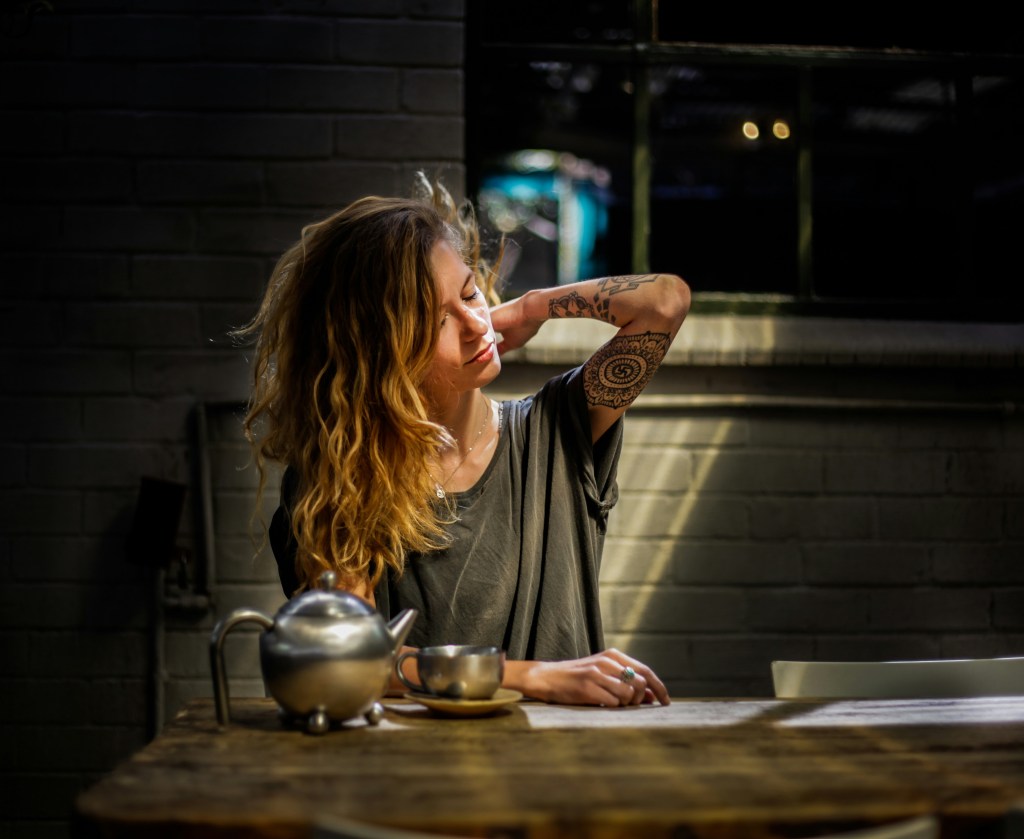 A highly stressed woman rubbing her neck while making tea. She is sitting at a nice wood table. 