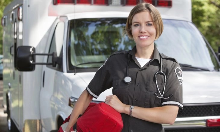 A smiling female paramedic holding her gear bag in front of an ambulance.