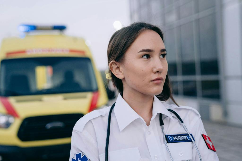 Female Paramedic looking to the left side of the screen with a yellow ambulance in the background. 