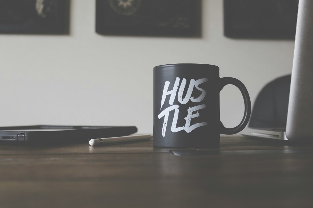 Here is a mug on a wooden table. The mug is black and says "Hustle" on it. 